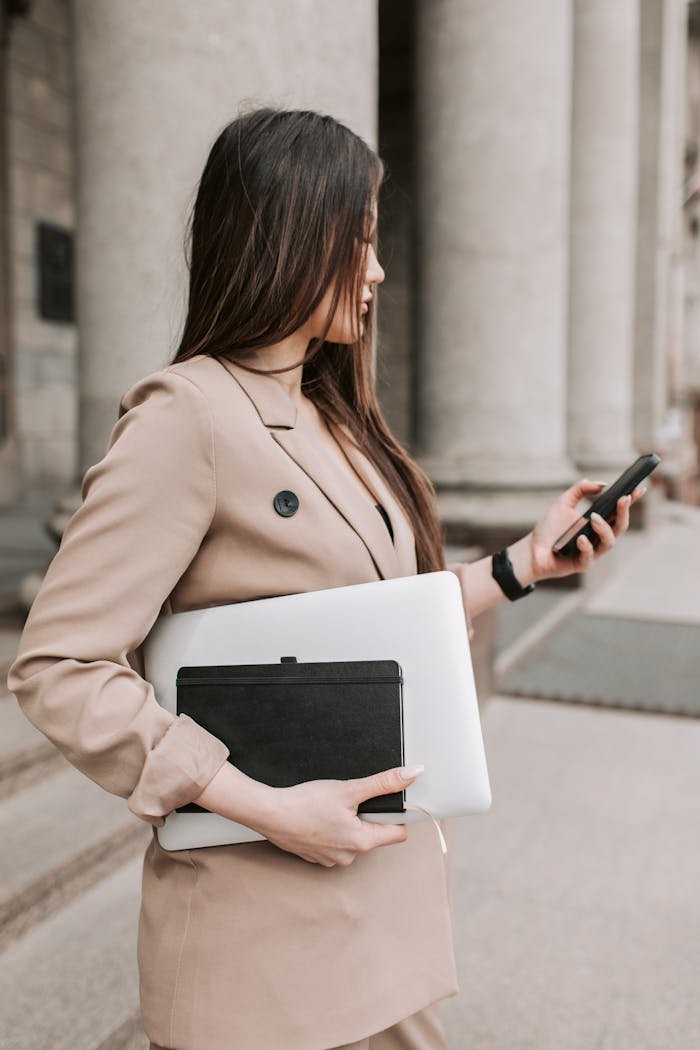 Confident young woman checking her phone while holding a laptop and notebook outside a building.